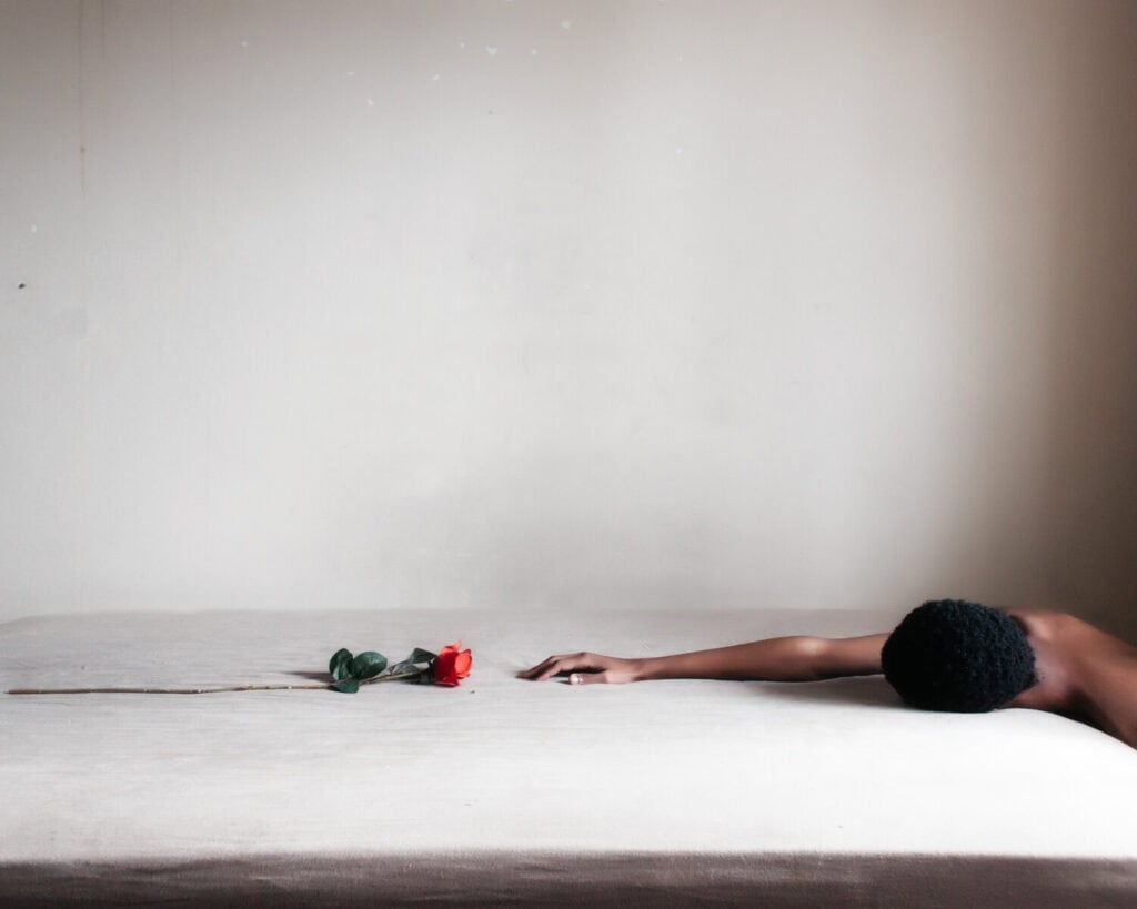 A person lying on a bed reaching towards a single rose, emphasizing themes of longing and tenderness.