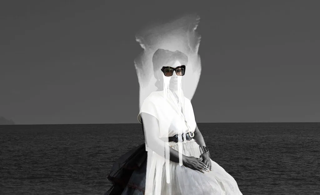 A woman in black sunglasses and layered clothing is seated against a background of the ocean and sky, creating a surreal photo composition.