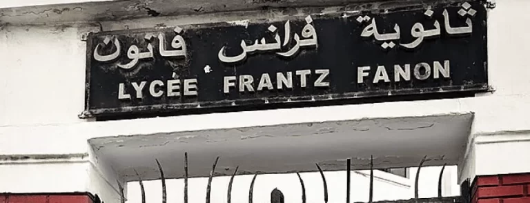 A black and white photo showing a street sign for Lycée Frantz Fanon with Arabic, French, and English text.