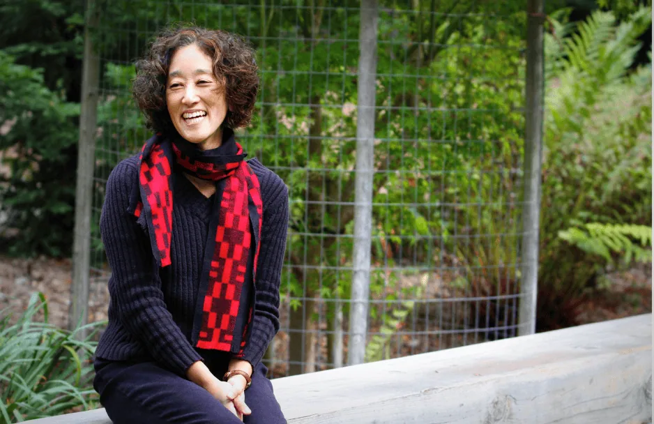 Karen Tei Yamashita seated outdoors with bushes in the background smiling/laughing.
