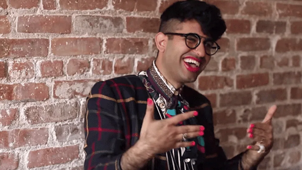 Alok Vaid-Menon with a big smile and hands in motion against an exposed brick wall.