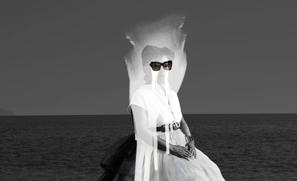 A woman in black sunglasses and layered clothing is seated against a background of the ocean and sky, creating a surreal photo composition.