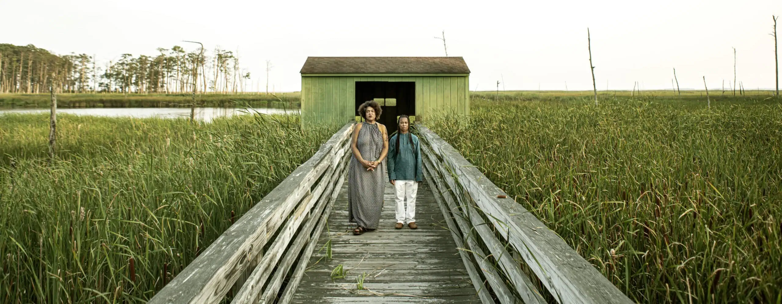 A woman and a girl walk hand in hand along a weathered wooden bridge over marshland grass, leading to a small green cabin.