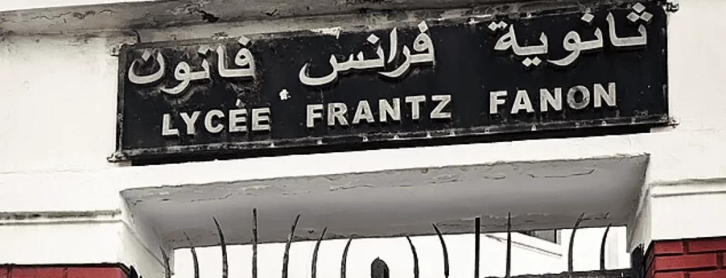 A black and white photo showing a street sign for Lycée Frantz Fanon with Arabic, French, and English text.